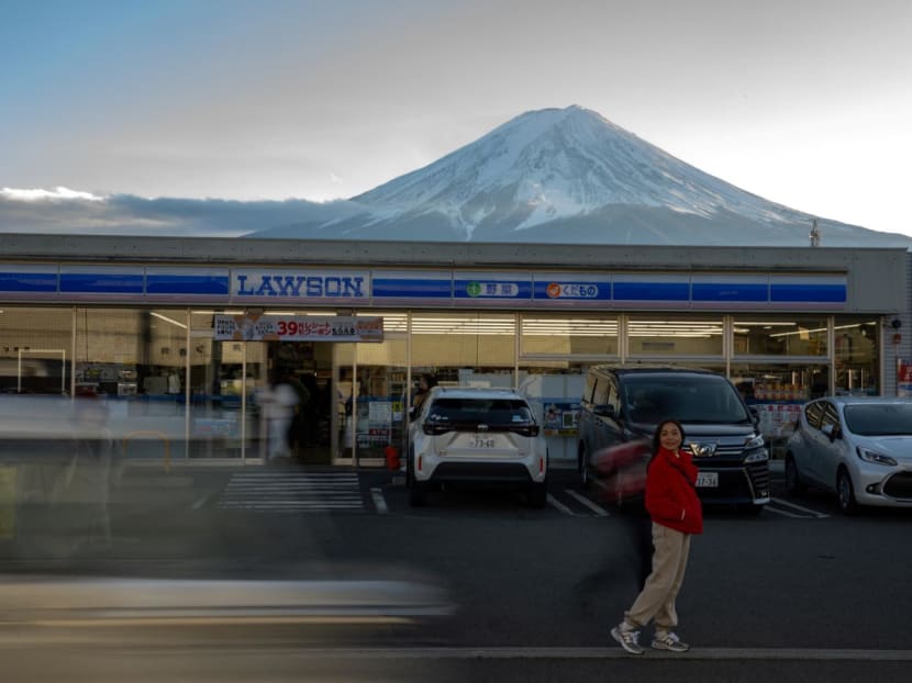 A tourist posing in front of a convenience store with Mount Fuji in the background, in the town of Fujikawaguchiko, Yamanashi prefecture on Jan 1, 2024. A tourist posing in front of a convenience store with Mount Fuji in the background, in the town of Fujikawaguchiko, Yamanashi prefecture on Jan 1, 2024.