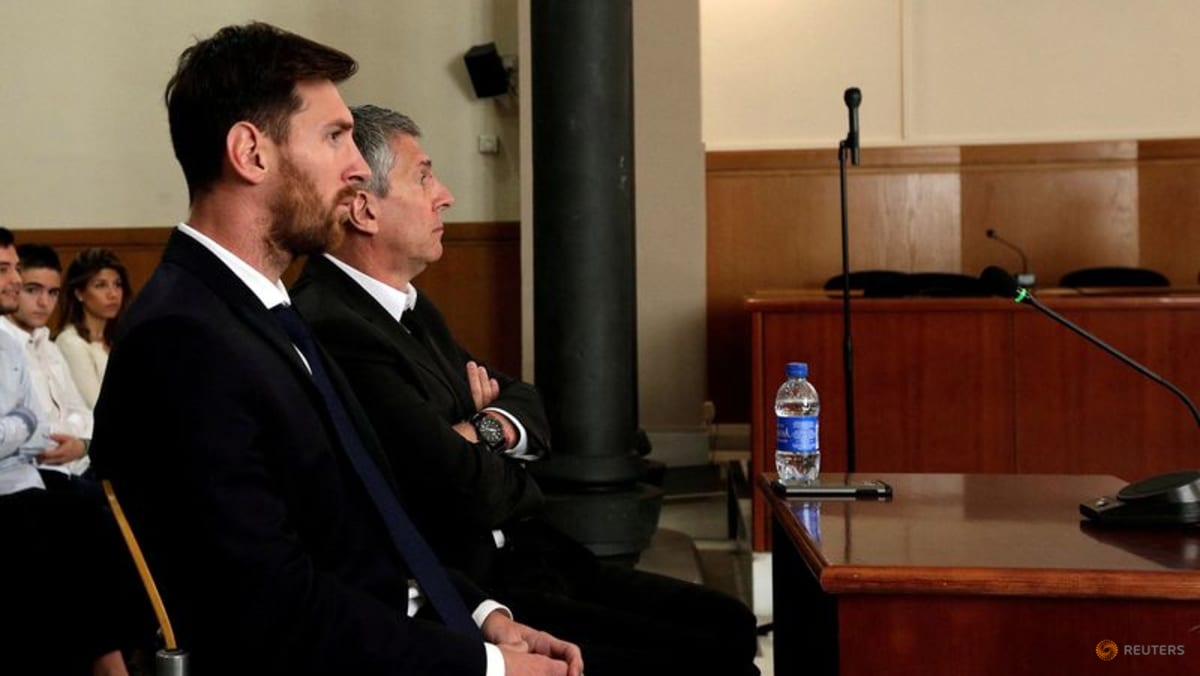 Two men in suits seated indoors in front of a wooden panel.