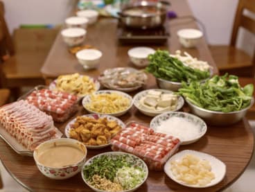 Fried and raw food items, along with peanut sauce for dipping, are set at a dining table, ready to be cooked in a hot pot of soup or broth for a communal meal.