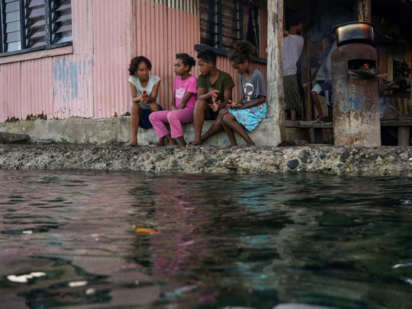 Children from a village in Fiji sitting outside their home next to a flooding sea wall at high tide. 