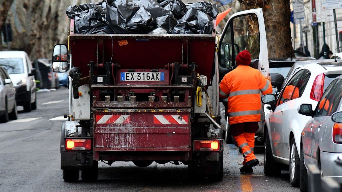 A worker in orange stands next to a red garbage truck loaded with black bags on a city street.