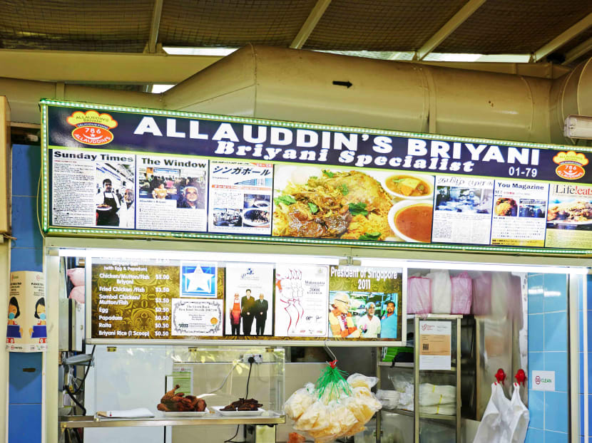 The image displays the storefront of Allauddin's Briyani, a "Briyani Specialist" stall numbered 01-79 that is adorned with numerous newspaper clippings and media features. Below the main signage, a detailed menu with prices is posted alongside a prominent photograph of the stall owners with the President of Singapore in 2011.